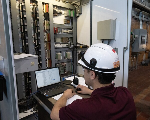 A man wearing a hard hat inspects electrical and instrumentation systems and records data on a laptop.