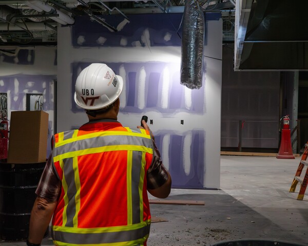 Inspector wearing a hard hat and vest shines a flashlight while walking through a partially completed building interior.