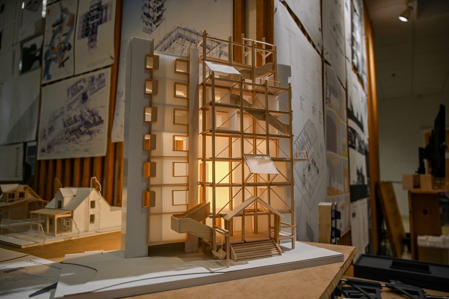 Student architectural model of a multi-story building exhibited in a studio space alongside architectural drawings and design concepts