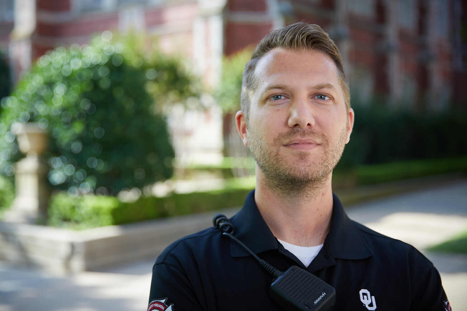 A person in a dark uniform with a radio microphone stands outdoors near greenery and a red‑brick building, including the word "staff" in all caps.