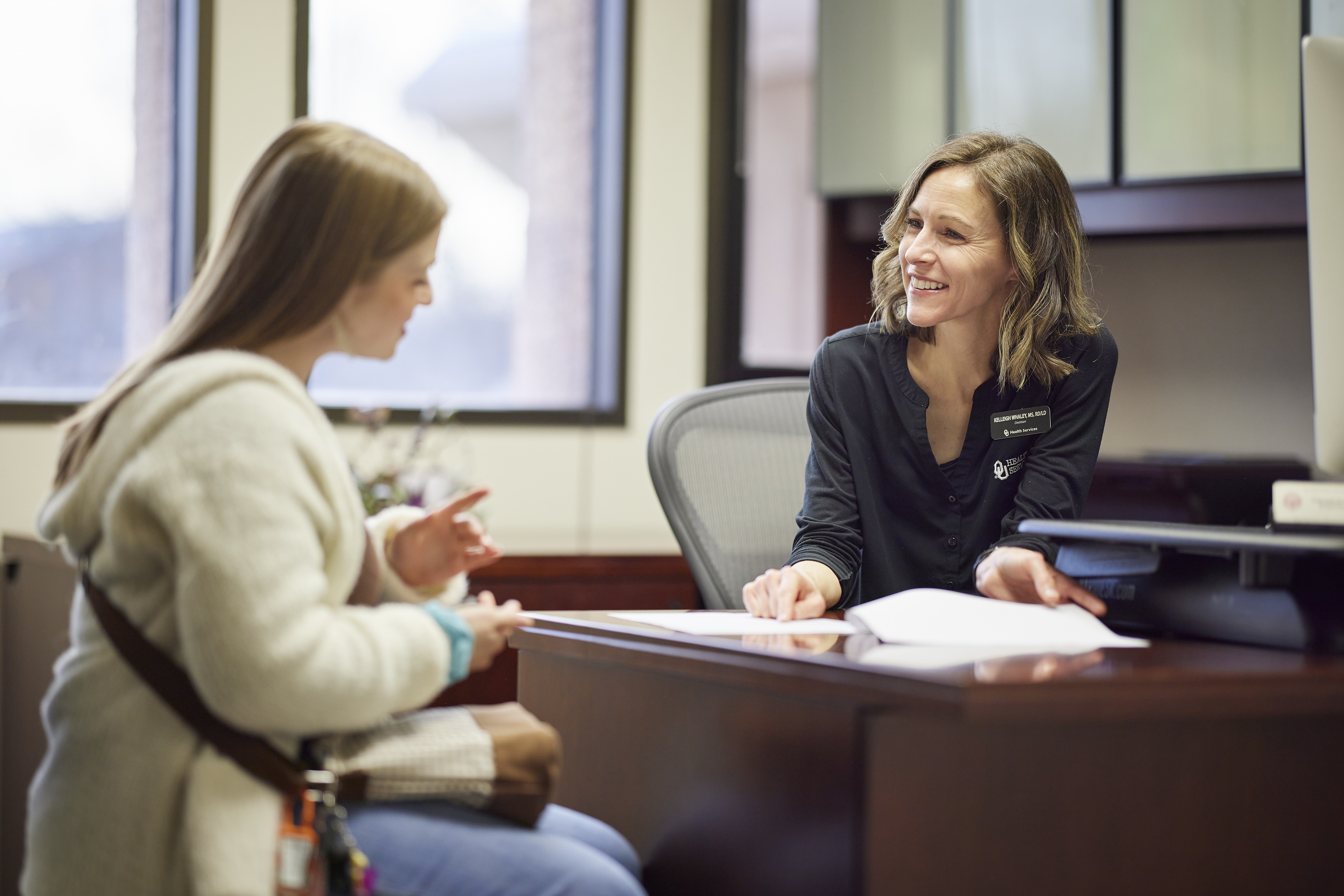 Two people sit across a desk in an office, reviewing papers together.